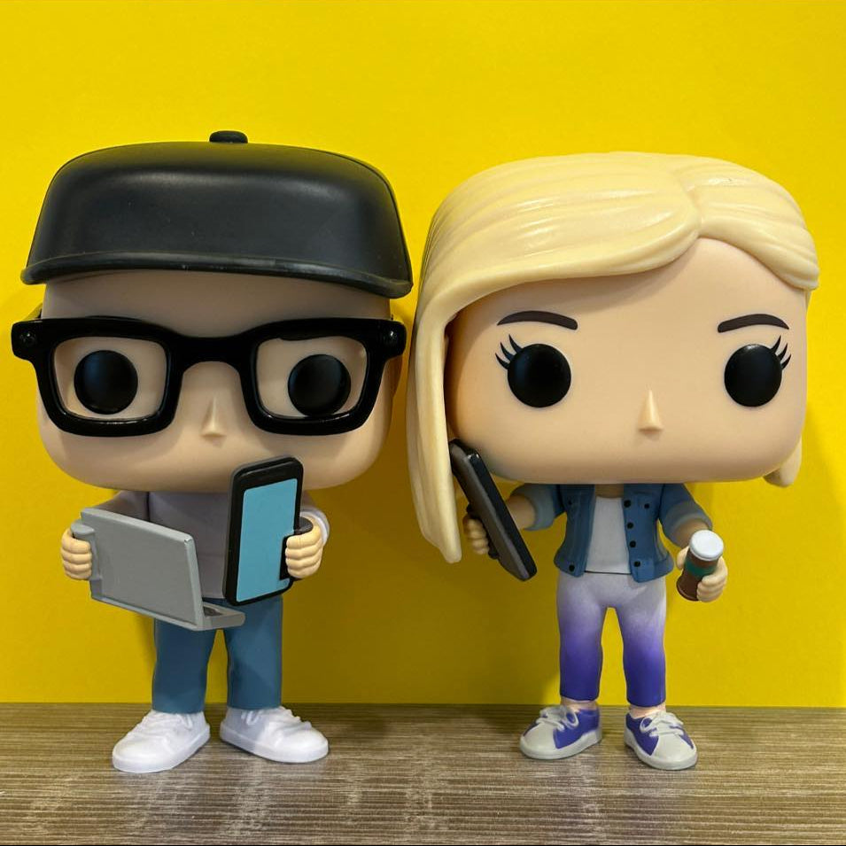 Two figurines, one with a cap and glasses holding a tablet, and the other with blonde hair holding a gun, against a yellow background.